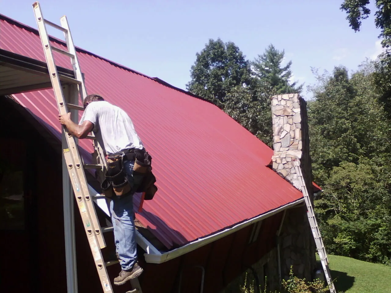 Roofer on a ladder beside a red metal roof and stone chimney — Soffit & Fascia Repair by Roofing Brothers in Royal Center